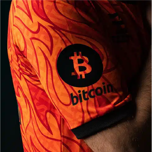 Un primo piano su una maglia arancione del Vesta Calcio con motivi intricati, mostrando il logo di Bitcoin, simboleggiando una partnership unica tra sport e criptovaluta.