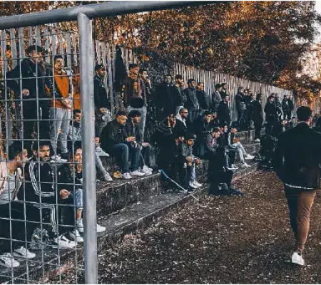 Una folla di tifosi assortiti osserva con attenzione una partita del Vesta Calcio dalle gradinate all'aperto, separate dal campo da una rete metallica.